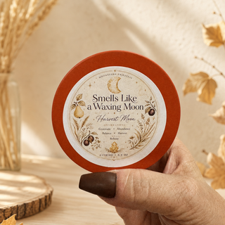 A hand with dark brown nail polish holds a round tin of body butter. The tin has a rust-orange rim and a cream-colored label that reads 'Smells Like a Waxing Moon - Harvest Moon' with illustrations of a crescent moon, pumpkin, and acorn.