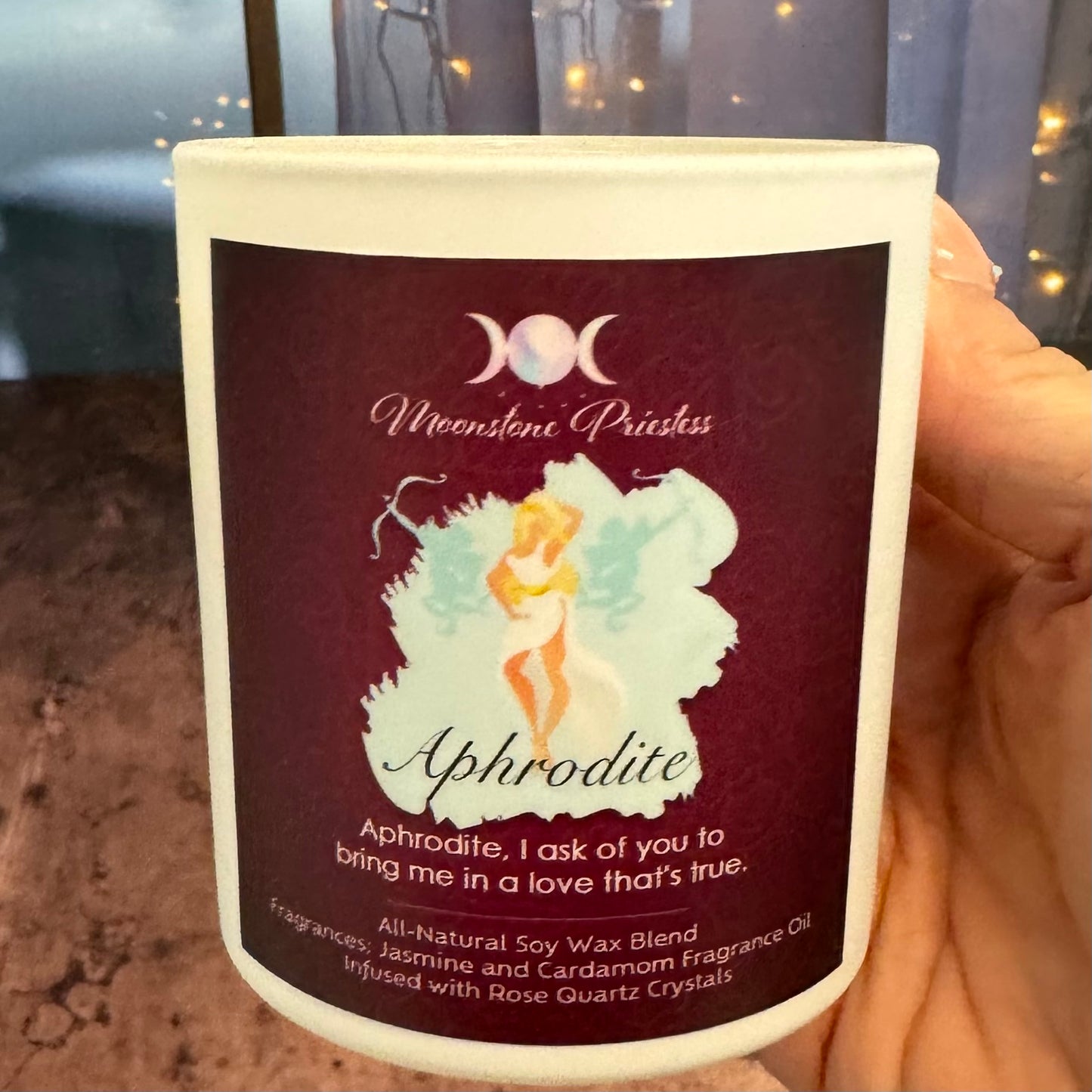 Candle with 'Aphrodite' label featuring a fairy design, held by a hand.