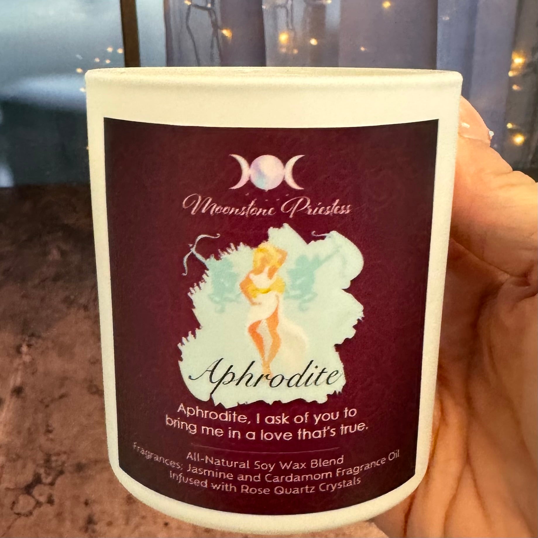 Candle with 'Aphrodite' label featuring a fairy design, held by a hand.