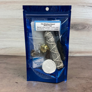 A blue resealable pouch containing a small ritual or herb bundle labeled 'The Shadow Guard White Sage,' with visible white sage bundles, a brass bell, a white powder circle, and a small blue card reading 'Vrinda Blue,' photographed against a light wood background.