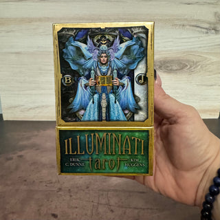 A hand holds an Illuminati Tarot deck, showing the gold-bordered card back with a blue and purple winged figure illustration and the green 'ILLUMINATI tarot' box front with creator names, against a light wood and dark brown background.
