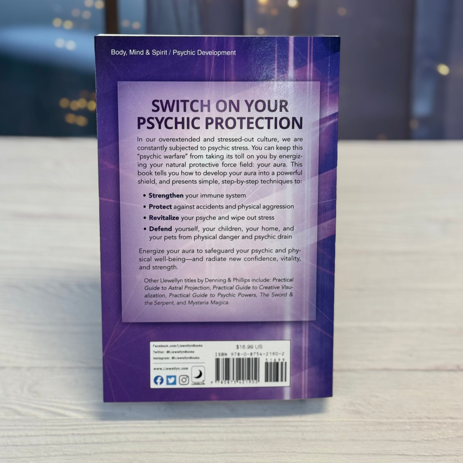 Back cover of a book titled 'Switch On Your Psychic Protection' on a wooden surface with blurred lights in the background.