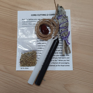 A cord cutting ritual kit laid out on a light wood tabletop, including a white printed instruction card, a clear plastic bag with dried herbs, a black and white wand, a bundle of white sage tied with purple string, a small bundle of brown herbs wrapped in twine, and a dark red oval stone.