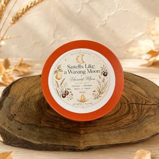 A round tin of citrus wax melts sits on a rustic wood slice. The tin has a bright orange rim and a cream label with botanical illustrations and the text 'Smells Like a Waxing Moon' and 'Harvest Moon'.
