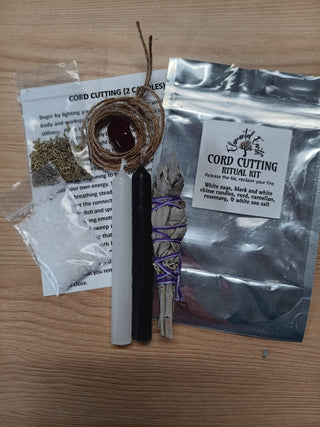 A cord cutting ritual kit laid out on a wooden surface, including a silver pouch labeled 'Cord Cutting Ritual Kit,' a white sage bundle wrapped in purple string, a black and white wand, a bundle of white sage with a brown cord, and a small packet of dried herbs.