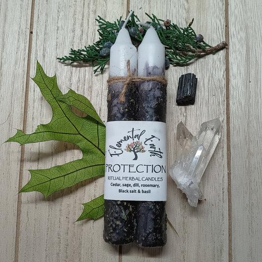 Two candles wrapped in black twine with a label, surrounded by greenery and crystals on a wooden surface.