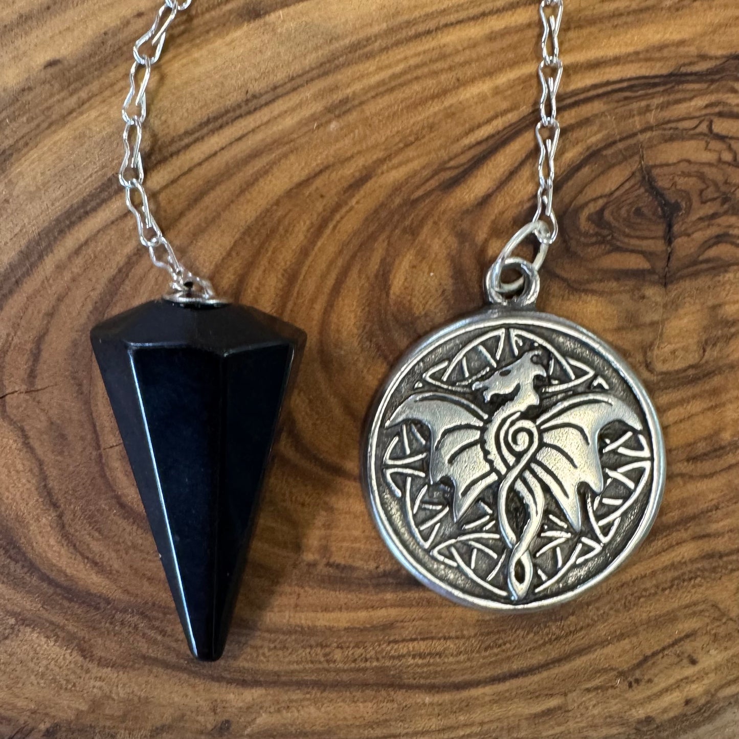 Black obsidian triangular pendulum and silver circular pendant with wolf design on a wooden surface