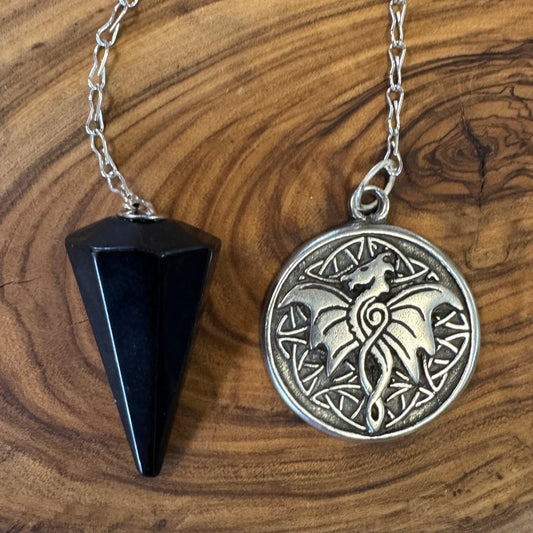 Black obsidian triangular pendulum and silver circular pendant with wolf design on a wooden surface