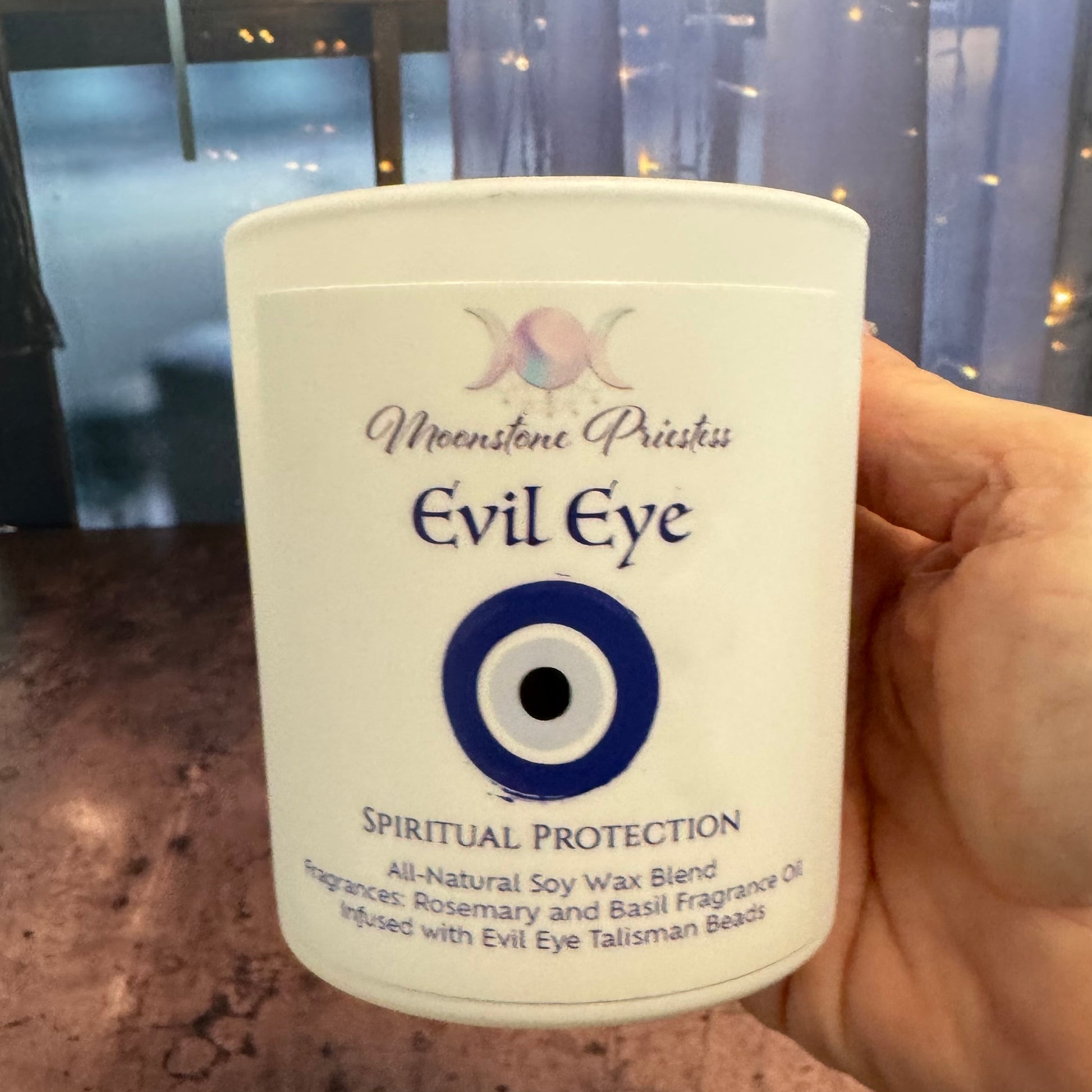 Hand holding a container labeled 'Evil Eye' with a blue and white design, against a blurred indoor background.