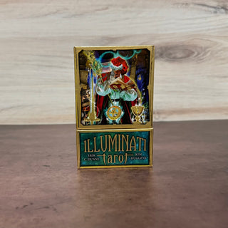 A boxed tarot card deck standing upright on a wooden surface against a light wood wall. The box has a gold border and a front panel showing an illustrated tarot figure with a red hat and a teal lower section reading 'ILLUMINATI tarot' with names 'KEITH DENNIS' and 'KIM HUGGINS'.