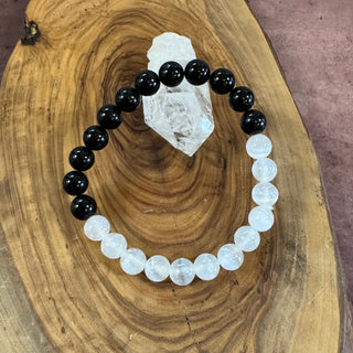 A beaded bracelet made of alternating glossy black and milky white round beads, arranged in an oval on a natural wood slice with a clear crystal cluster behind it.