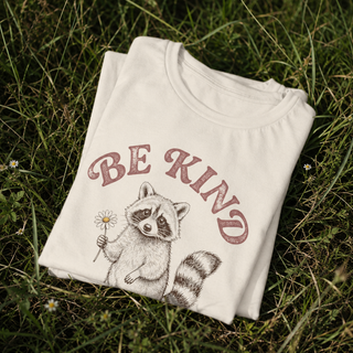 Folded white t-shirt with a raccoon graphic and 'BE KIND' text on grass