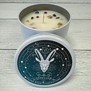 A white candle with a label featuring a Capricorn sign and zodiac symbols, surrounded by decorative stones. The candle is placed on a wooden surface.