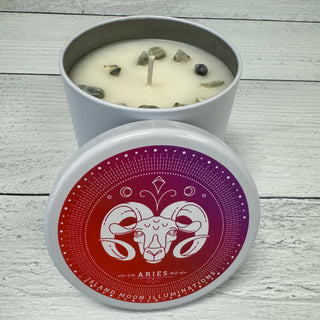 An Aries Zodiac themed candle with eucalyptus, vanilla, and mint fragrance, and serpentine crystals inside. The candle is placed on a wooden surface with a decorative label featuring the Aries sign and zodiac elements.
