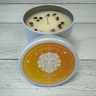 A candle with a label indicating it is a Leo Zodiac Immersion Candle, with orange and clove fragrances, and tiger's eye crystals. The candle is placed on a wooden surface.