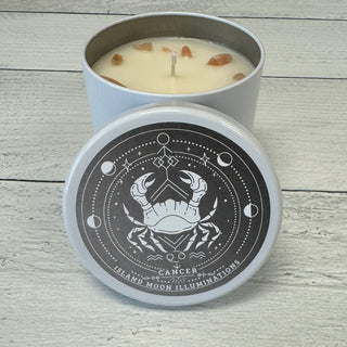 A candle with a label indicating it is a Cancer Zodiac Candle, with vanilla and buttercream fragrances and peach moonstone crystals. The candle is placed inside a white box with a design that includes a crab symbol, moon, and stars.