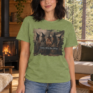 A woman wearing a heather green graphic t-shirt with a vintage-style print of four witches and the text 'Girls Will Be Girls.' in a cozy, wood-paneled room.