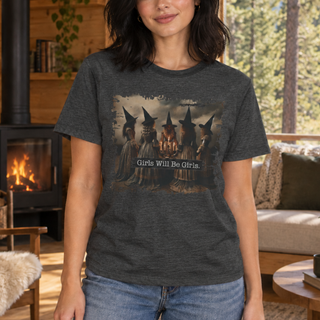 A woman wearing a dark heather gray graphic t-shirt with a vintage-style illustration of four witches and the text 'Girls Will Be Girls.' in a cozy, wood-paneled room.