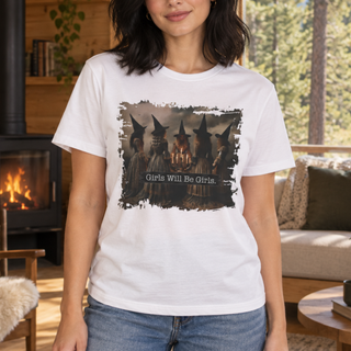 A woman wearing a white graphic t-shirt with a dark, distressed print of witch girls around a campfire and the text 'Girls Will Be Girls.' in a cozy, sunlit living room with a fireplace in the background.