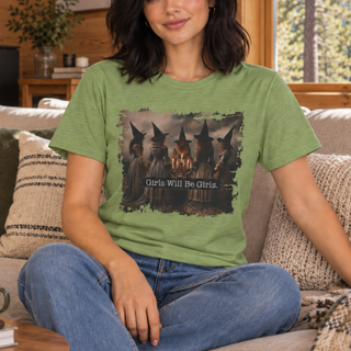 A woman sitting on a couch wearing a sage green graphic t-shirt with a dark illustration of three witches and the text 'Girls Will Be Girls'.