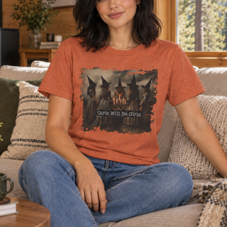 A woman sitting on a couch wearing a heathered rust-orange graphic t-shirt with a distressed print of witches and the text 'Girls Will Be Girls'.