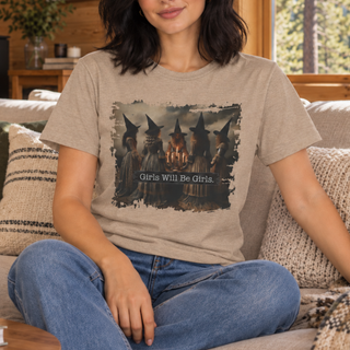 A woman sitting on a couch wearing a beige graphic t-shirt with a dark illustration of four witches and the text 'Girls Will Be Girls.' She is also wearing blue jeans.