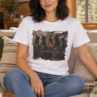 A woman sitting on a couch wearing a white graphic t-shirt with a distressed black-and-gray print of witches and the text 'Girls Will Be Girls'.