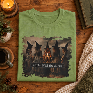 A folded sage green t-shirt with a graphic of four witches holding candles and the text 'Girls Will Be Girls.' displayed on a rustic wooden table surrounded by cozy lifestyle props like a candle, coffee, pinecones, and a knit blanket.