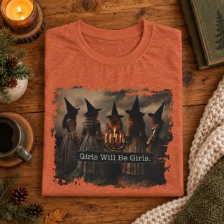 A folded heathered orange t-shirt with a graphic of four witches around a candle and the text 'Girls Will Be Girls.' displayed on a rustic wooden table with cozy lifestyle props like a candle, coffee, pinecones, and a knit blanket.