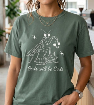 Person wearing a blue spruce green t-shirt with Girls Will Be Girls praying mantis graphic in a casual outdoor setting.
