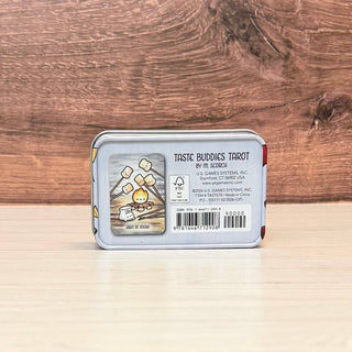A square metal tin shown from the back, featuring a label for 'Taste Buddies Tarot by Mr. Scrooge' with a cartoon illustration of a yellow duck and marshmallows, placed on a light wood surface against a darker wood background.