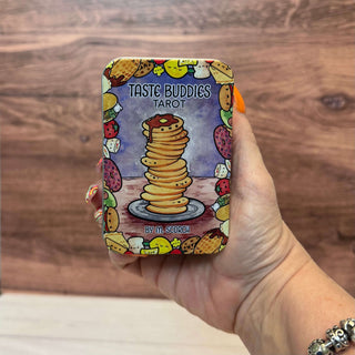 A hand holds a square tin of Taste Buddies Tarot against a wooden background. The tin features a colorful illustration of a tall stack of pancakes on a plate, surrounded by food-themed doodles and the text 'TASTE BUDDIES TAROT' and 'BY M.