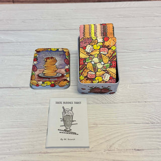 Taste Buddies Tarot deck displayed on a light wood surface, including a tin box with illustrated cards, a smaller tin lid, and a card back sheet reading 'TASTE BUDIOKS TAROT By M. Scorch'.