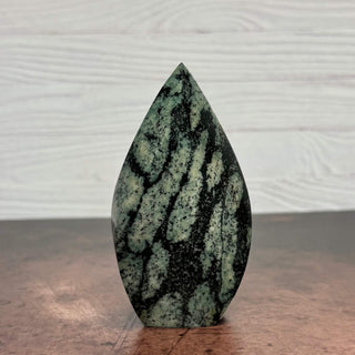 A polished teardrop-shaped green stone with dark black and gray veining, standing upright on a wooden surface against a light gray wood-paneled background.