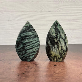 Two polished teardrop-shaped stone sculptures with green and black marbled patterns, displayed on a dark wooden surface against a light wood paneled wall.