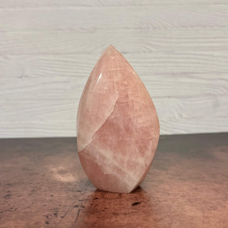 A polished pink rose quartz crystal tower with natural striations, standing upright on a dark wooden surface against a light gray wood-paneled background.