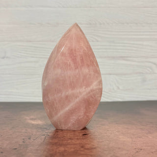 A polished pink rose quartz crystal point sculpture standing upright on a dark wooden surface against a light gray wood-paneled wall.