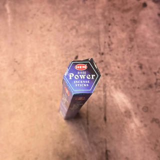 A hexagonal box of HEM Divine Power Incense Sticks standing upright on a textured brown surface, viewed from a high-angle perspective.