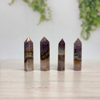 Four polished natural amethyst crystal points of varying heights standing upright on a light wooden surface, with a soft-focus white wall and green foliage in the background.