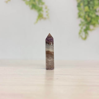 A polished natural amethyst crystal point tower standing upright on a light wooden surface, with a soft-focus background of green foliage and a white wall.