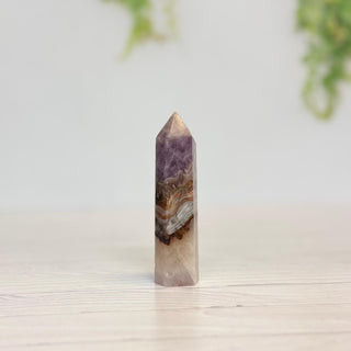 A natural quartz crystal point featuring purple amethyst and brown agate bands, standing upright on a light wooden surface against a soft, blurred background.