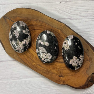 Three polished black jasper stones with pink and white matrix patterns arranged on a natural wood serving board over a white wood plank background.