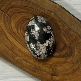 A polished black and white spotted stone, resembling a jasper egg, displayed on a rustic live-edge wooden board.