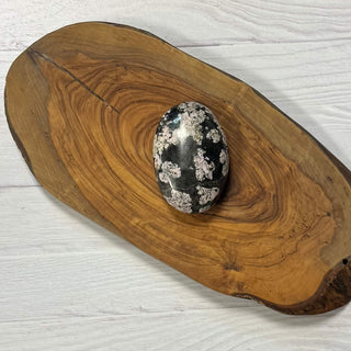 A polished black and white mottled stone palm resembling a jade palm, displayed on a rustic live-edge wooden serving board over a white painted wood background.