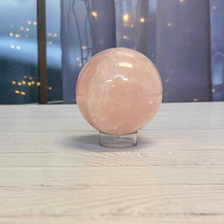 A polished pink rose quartz crystal sphere resting on a clear acrylic display stand, placed on a light-colored wooden tabletop with a softly blurred background.