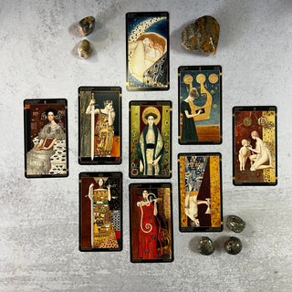 A collection of tarot cards from the Golden Tarot of Klimt deck, laid out on a grey surface, accompanied by stones and a booklet.