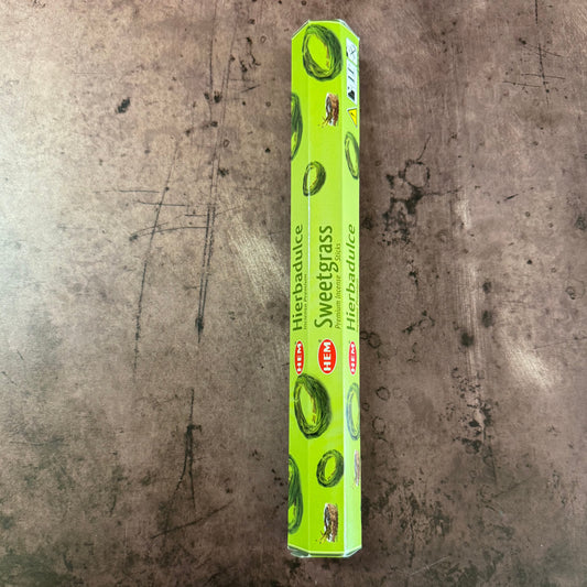A green box of Sweetgrass Hem Hexagonal incense with branding and images of sweetgrass on it, placed on a textured metallic surface.