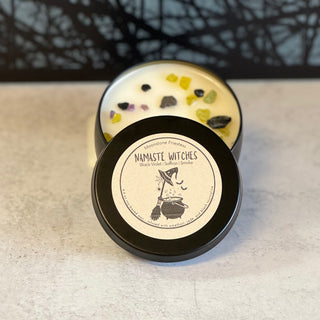 A black container with a lid, featuring a white candle inside, adorned with colorful crystals at the base and a label on the lid with 'Namaste Witches' 