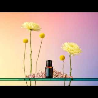 A small amber glass essential oil bottle with a black cap stands on a clear glass shelf, surrounded by pale pink filler flowers and yellow daisy-like blooms against a soft pink-to-lavender gradient background.