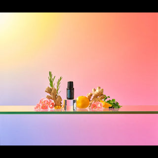 A small black essential oil bottle with a blue label stands on a reflective surface, styled with ginger roots, pink flowers, and citrus slices against a pastel gradient background.